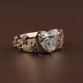 May include: A gold ring with a heart-shaped diamond. The ring is decorated with small pink gemstones and gold flower details. The wide, polished band adds to the ring's elegant design. A striking piece of jewellery.