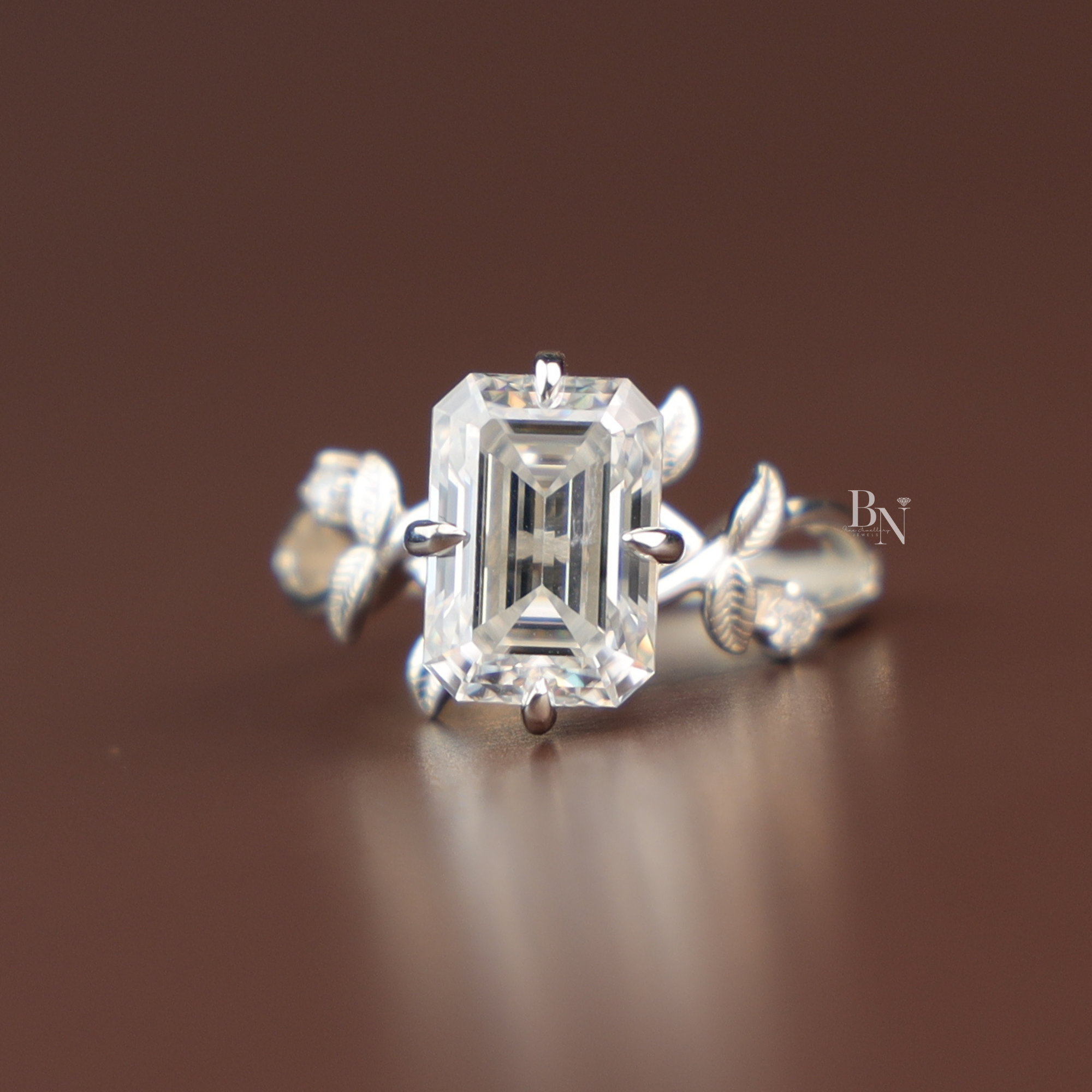 A magnificent 3ct emerald cut lab grown diamond engagement ring in polished white gold, featuring an intricate vine-inspired shank with delicate leaf details, hidden halo, and sparkling accents