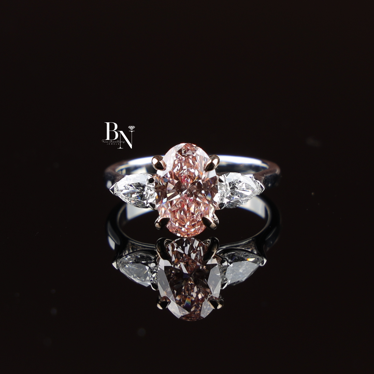 Three-stone engagement ring featuring a 2.2 carat fancy pink oval lab-grown diamond center stone in rose gold claw prongs, flanked by two colorless pear-cut side diamonds on a polished white gold band.
