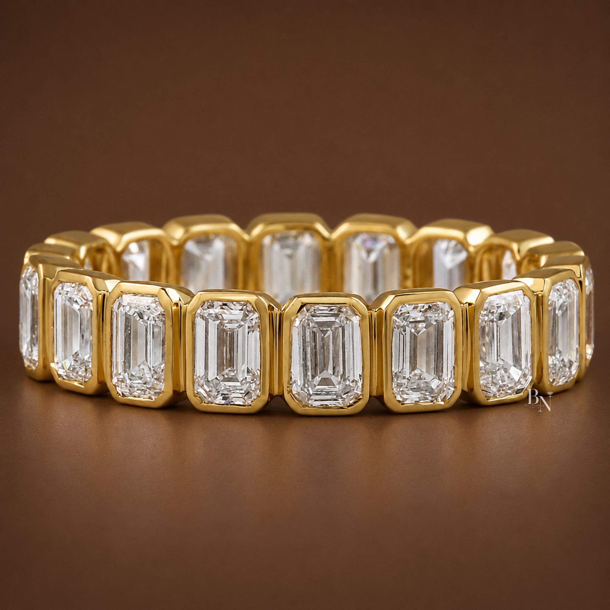 Angled front view of a yellow gold eternity band with bezel-set emerald cut lab diamonds resting on a dark brown background.