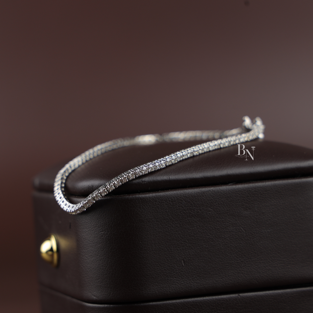 A classic silver-tone tennis bracelet resting on a dark brown textured leather box, showcasing a continuous line of round-cut diamonds in a four-prong setting.