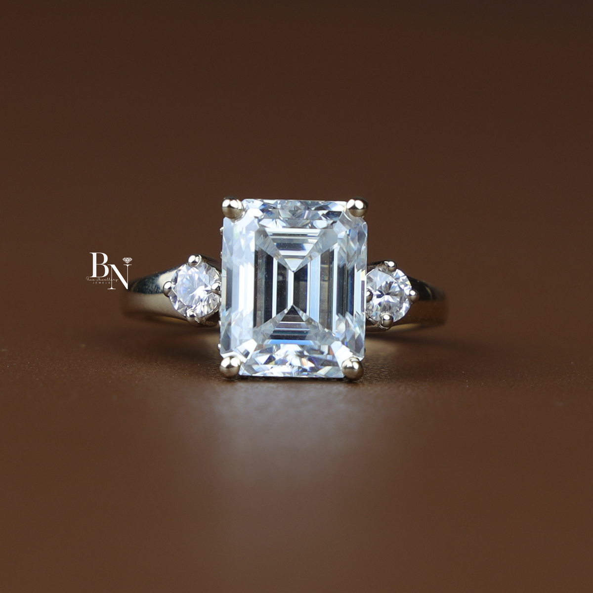A 5 carat emerald cut moissanite engagement ring in yellow gold, featuring a classic three-stone design with round side stones and a hidden halo.