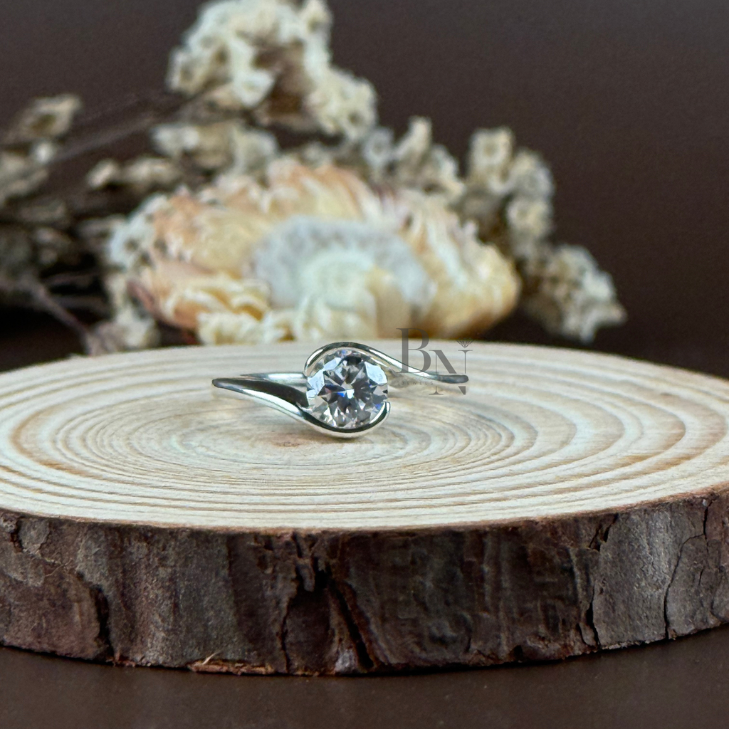 1 carat round brilliant cut lab grown diamond securely held in a modern bypass bezel swirl setting. The 14k white gold band features a high-polish finish and an asymmetrical wrap-around design shown on a hand and a wooden display.