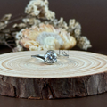 1 carat round brilliant cut lab grown diamond securely held in a modern bypass bezel swirl setting. The 14k white gold band features a high-polish finish and an asymmetrical wrap-around design shown on a hand and a wooden display.