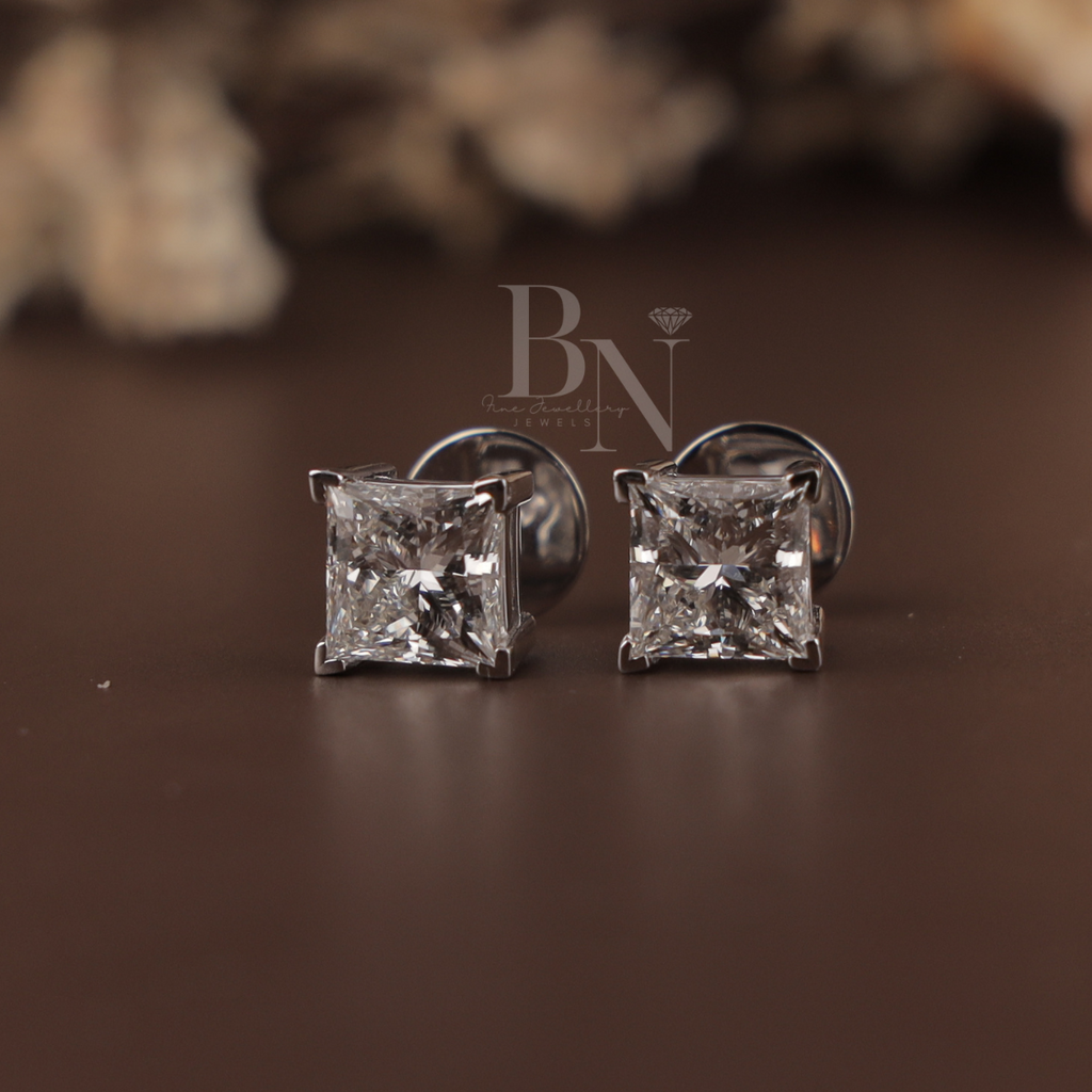 May include: Two square-cut diamond stud earrings set in silver metal, presented in a black jewellery box with a velvet lining. The diamonds are shown sparkling. The earrings are a classic design.