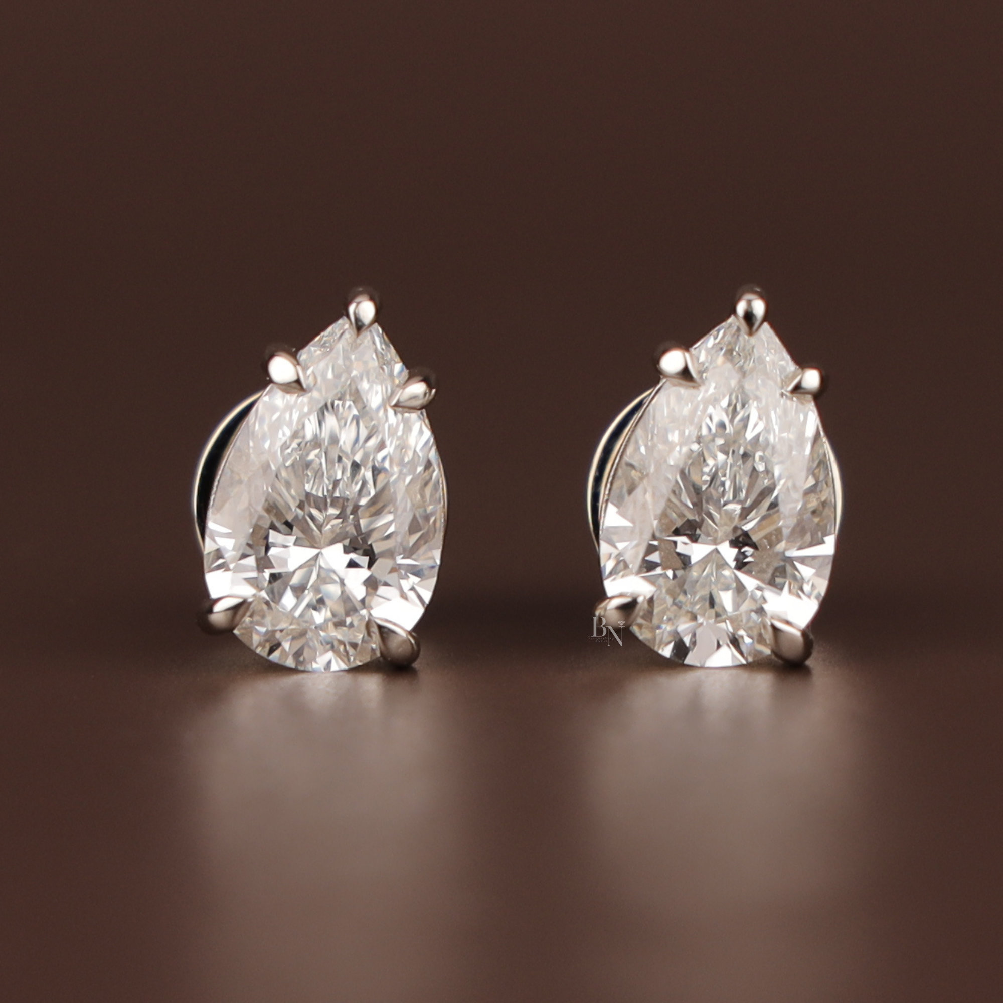 May include: A pair of pear-cut diamond stud earrings. Each earring showcases a clear, faceted diamond set in a silver-coloured metal. The diamonds are the focal point, reflecting light against a dark brown background.