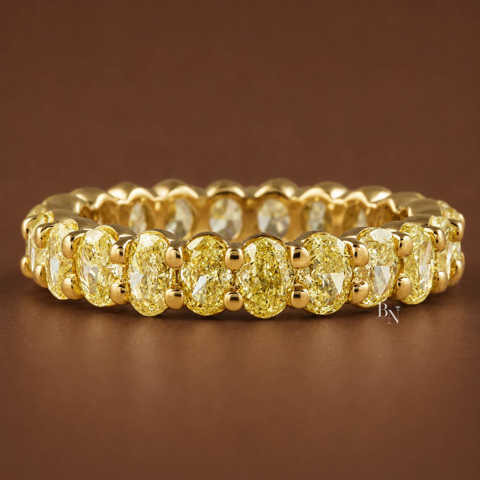 Angled front view of an 18K yellow gold eternity band with fancy yellow oval diamonds resting on a dark brown background.