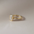 Macro view of a 2 carat emerald cut lab grown diamond securely held in a modern 14k yellow gold bezel setting with a polished rounded solitaire band and sleek low profile, showcasing step-cut clarity and sophisticated bridal design.