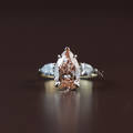 Three stone engagement ring featuring a 3 carat fancy pink pear cut lab grown diamond center stone with protective claw prongs, flanked by two white pear cut side diamonds on a tapered 14k yellow gold band.