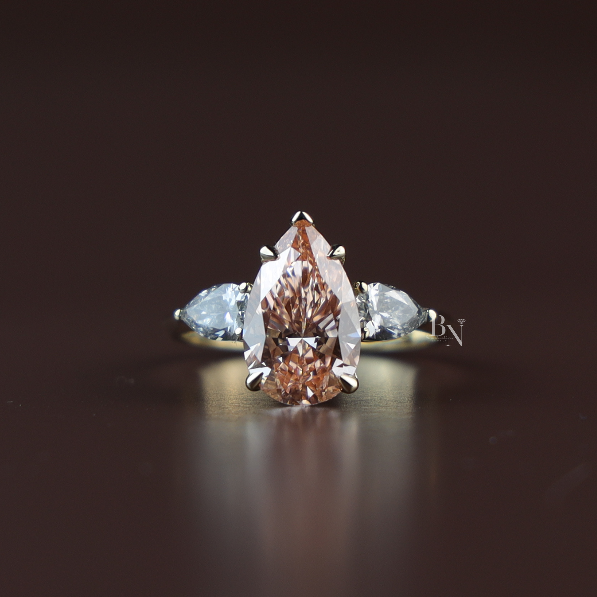 Three stone engagement ring featuring a 3 carat fancy pink pear cut lab grown diamond center stone with protective claw prongs, flanked by two white pear cut side diamonds on a tapered 14k yellow gold band.
