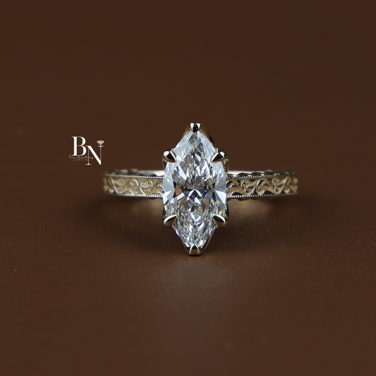 Side profile of a gold-tone engagement ring on a hand, featuring a marquise-cut diamond and an intricately engraved filigree band.