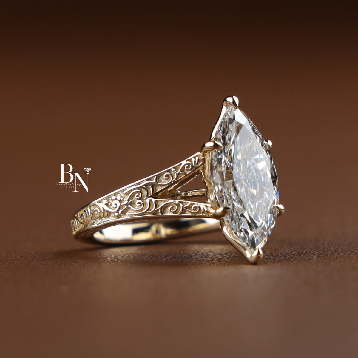 A 3 carat marquise-cut lab-grown diamond solitaire engagement ring with an engraved yellow gold filigree band held in black gloved fingers.