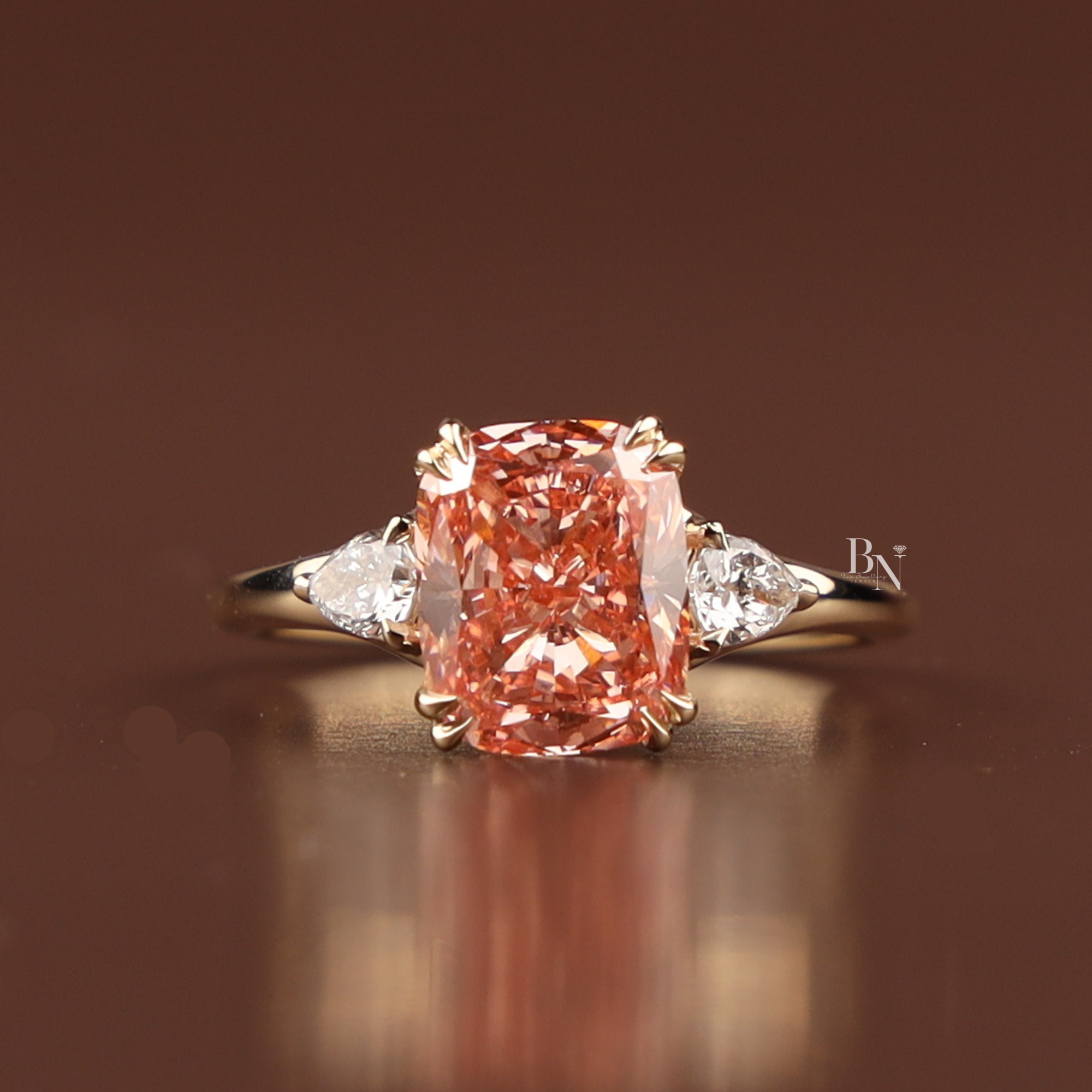 A 3 carat fancy pink cushion cut lab grown diamond engagement ring in yellow gold featuring pear shaped white diamond side accents and a double claw prong setting shown from multiple side angles and worn on a finger.