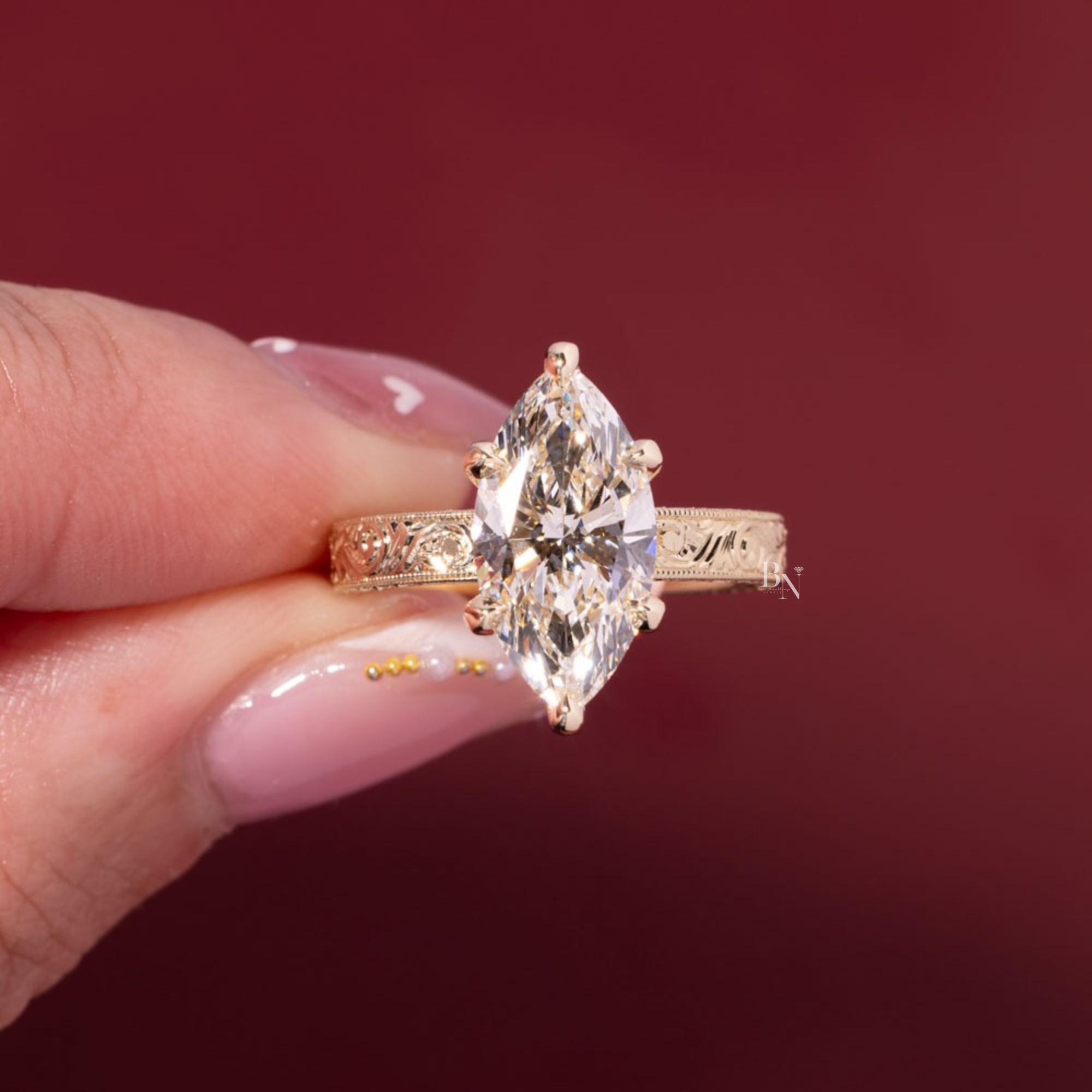 May include: A gold engagement ring with a marquise-cut diamond. The ring has an ornate, engraved band. The diamond is set in a prong setting. The ring is on a person's finger.