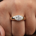 Macro view of a 2.50 carat oval lab grown diamond set horizontally in a 14k yellow gold half-bezel signet engagement ring with a thick polished tapered band.