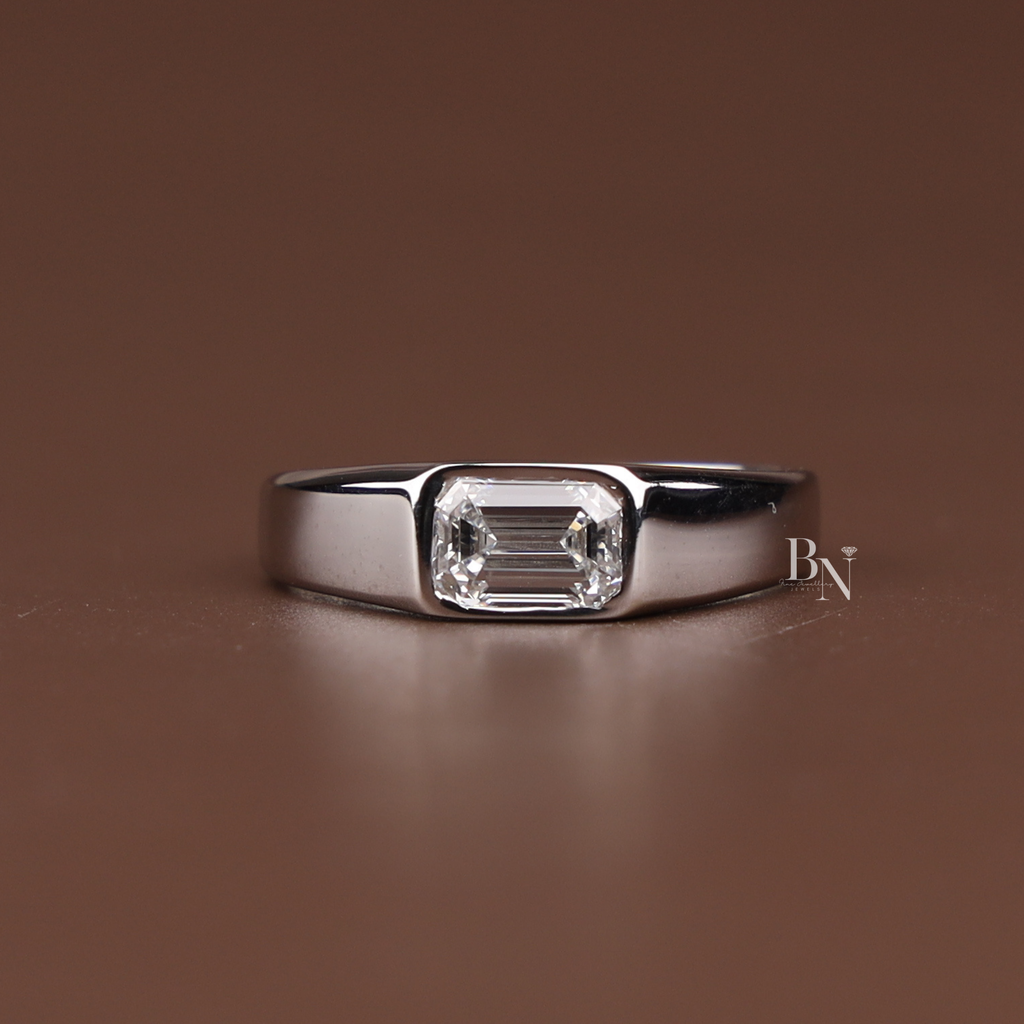 May include: A silver ring featuring a rectangular, clear gemstone. The gemstone is set in a bezel setting, and the band is thick and polished. The ring is displayed on a brown surface.
